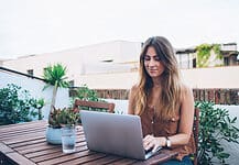 Cheerful woman with wristlet watch browsing laptop sitting on wooden chair on terrace while looking at screen in thoughts and working